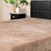 A close-up of a bed with a soft, light brown blanket and a cozy Teddy fleece hoeslaken - mink. In the background, there's a black nightstand with a plant and decor item, plus a black upholstered headboard.